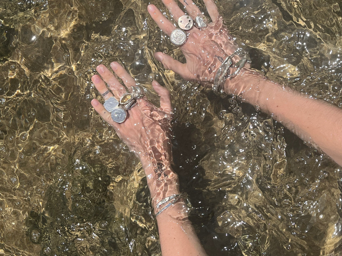 Two hands reaching into shallow, sunlit water while wearing multiple silver rings and bracelets.